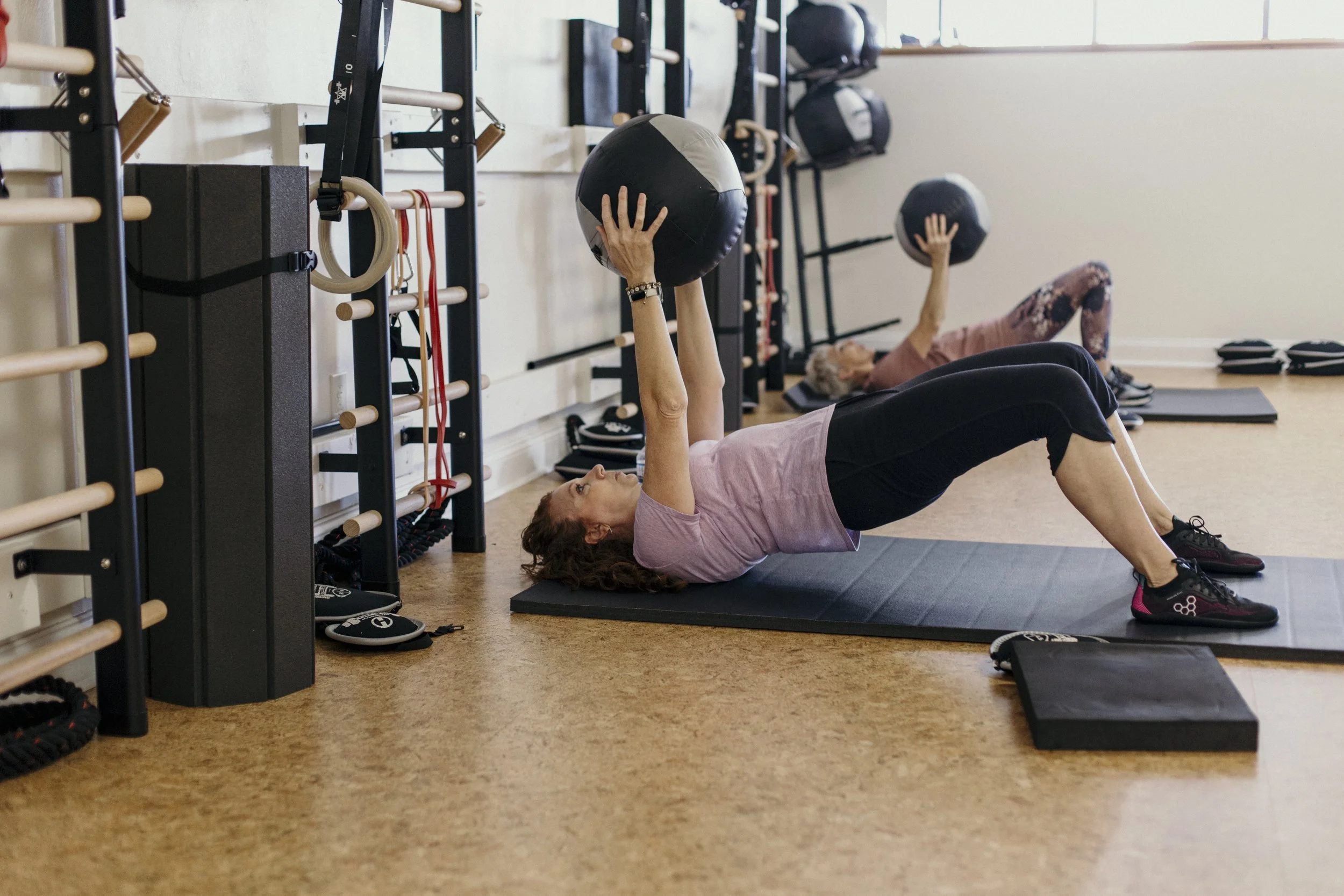 Two women performing glute bridges while holding medicine balls during a personal training session.