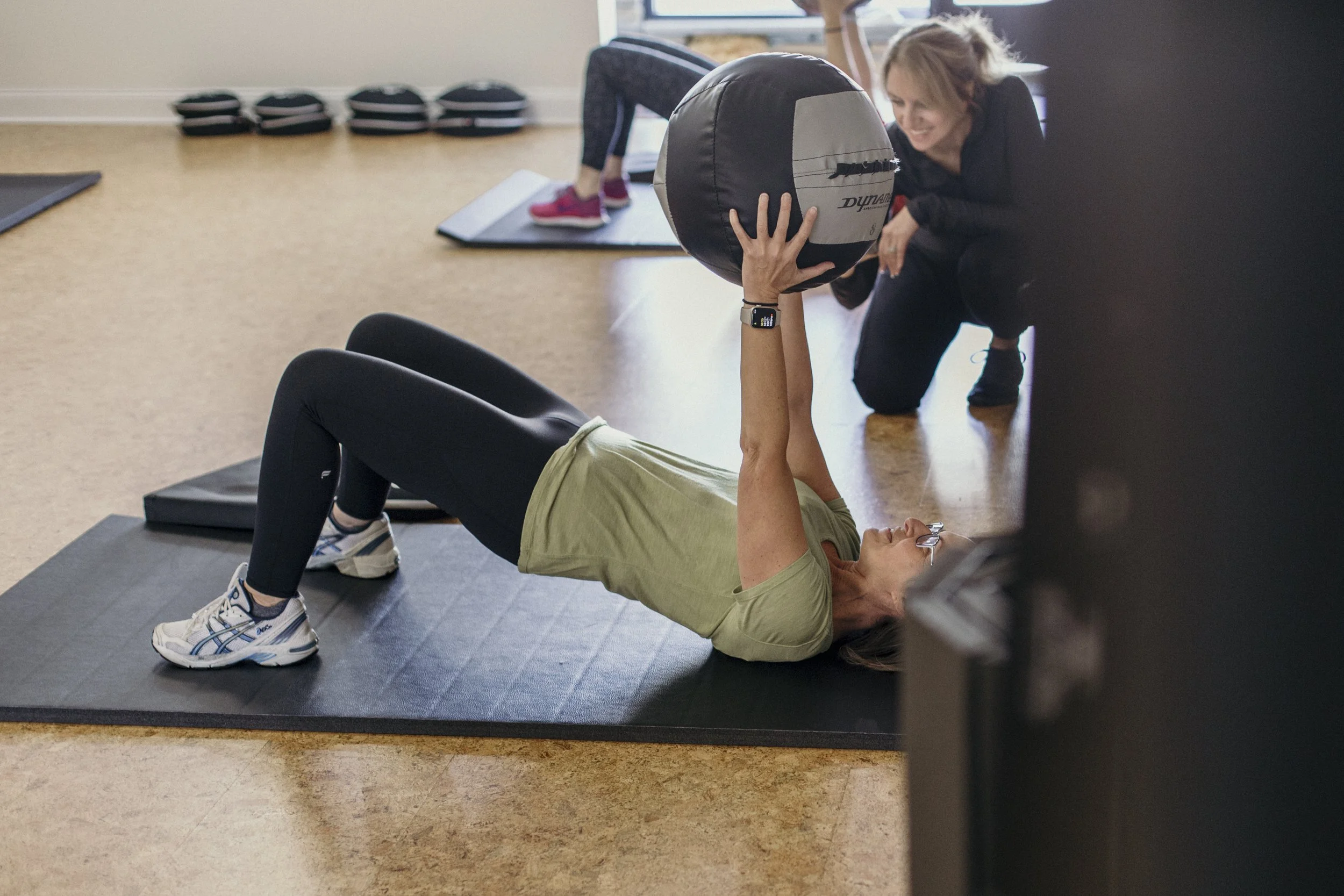 Woman performing a bridge exercise while holding a medicine ball as a personal trainer provides guidance.