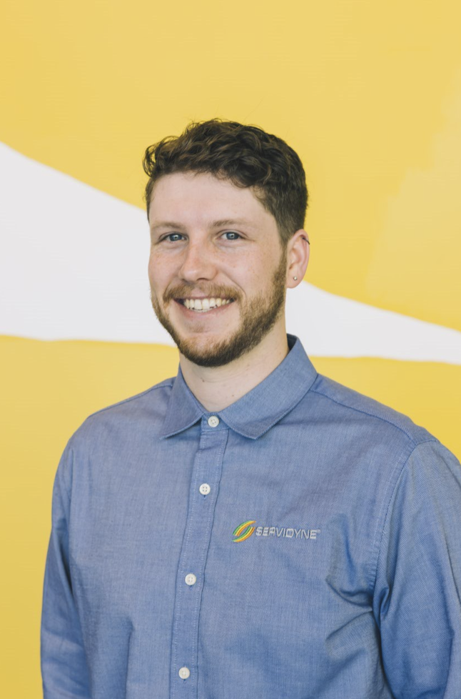 A young man with short, curly brown hair and a beard, smiling, wearing a blue button-up shirt with the SeavidYNE logo, standing in front of a yellow background.