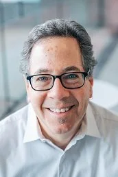 Close-up of a middle-aged man with dark hair, glasses, and a white shirt smiling in an office setting.