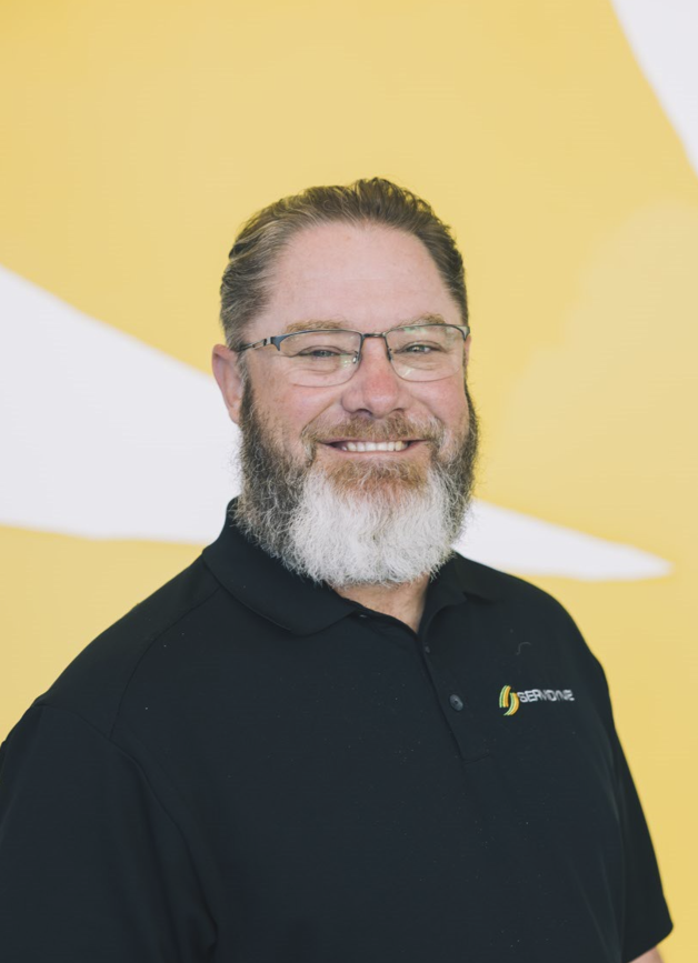 A smiling man with glasses and a beard, wearing a black collared shirt with a logo, standing in front of a yellow background.