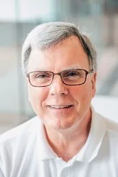 Smiling middle-aged man with gray hair and glasses, wearing a white shirt, in an office setting.
