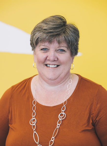 A woman with short gray hair, wearing a rust-colored top, silver jewelry, and smiling, standing against a yellow background.