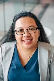 Portrait of a woman with glasses, wearing blue scrubs and a gray blazer, smiling in an indoor setting.