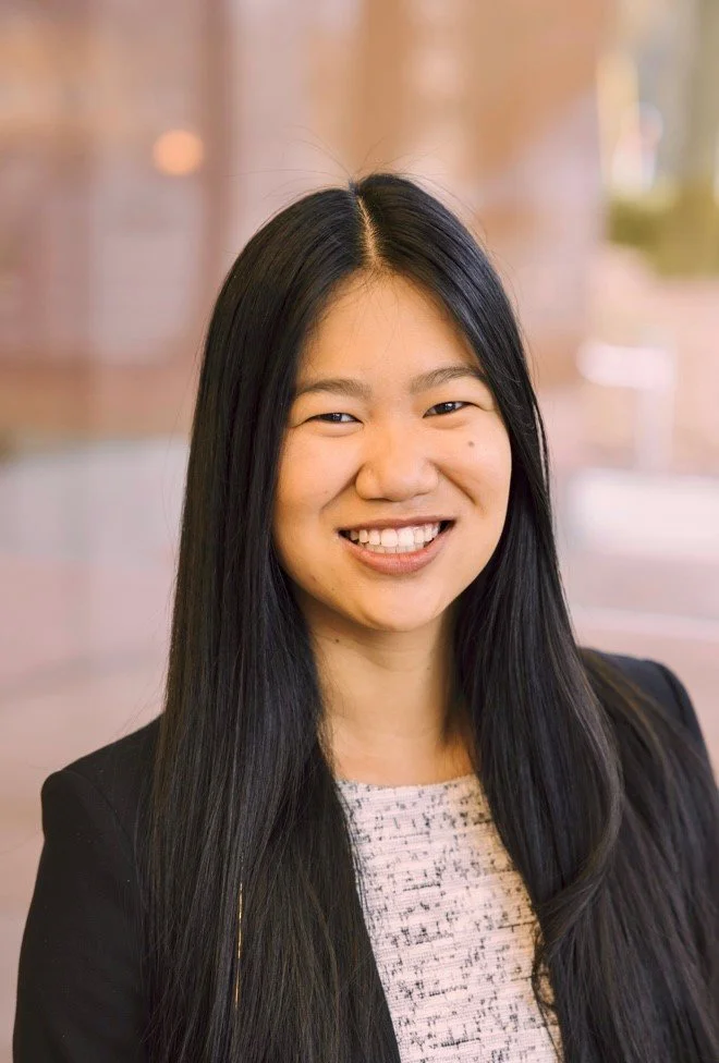 A portrait of a young woman with long black hair, smiling, wearing a black blazer over a light-colored top with a textured pattern, standing indoors with a blurred background.