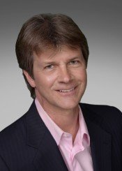 Portrait of a man with brown hair, wearing a dark blazer and pink shirt, smiling against a gray background.