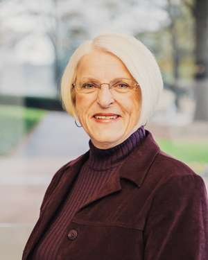 A smiling elderly woman with short white hair, wearing glasses, a maroon turtleneck sweater, and a dark jacket, standing outdoors with trees and a walkway in the background.