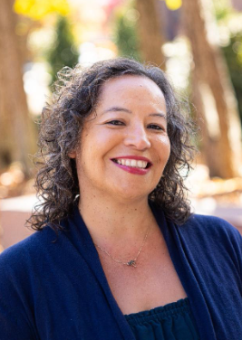 A woman with curly brown hair smiling outdoors in a forested area, wearing a navy blue top and a necklace.