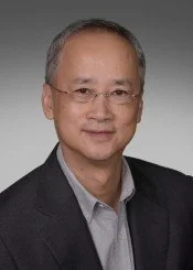 Portrait of a middle-aged man with short gray hair and glasses, wearing a dark blazer and light shirt against a gray background.