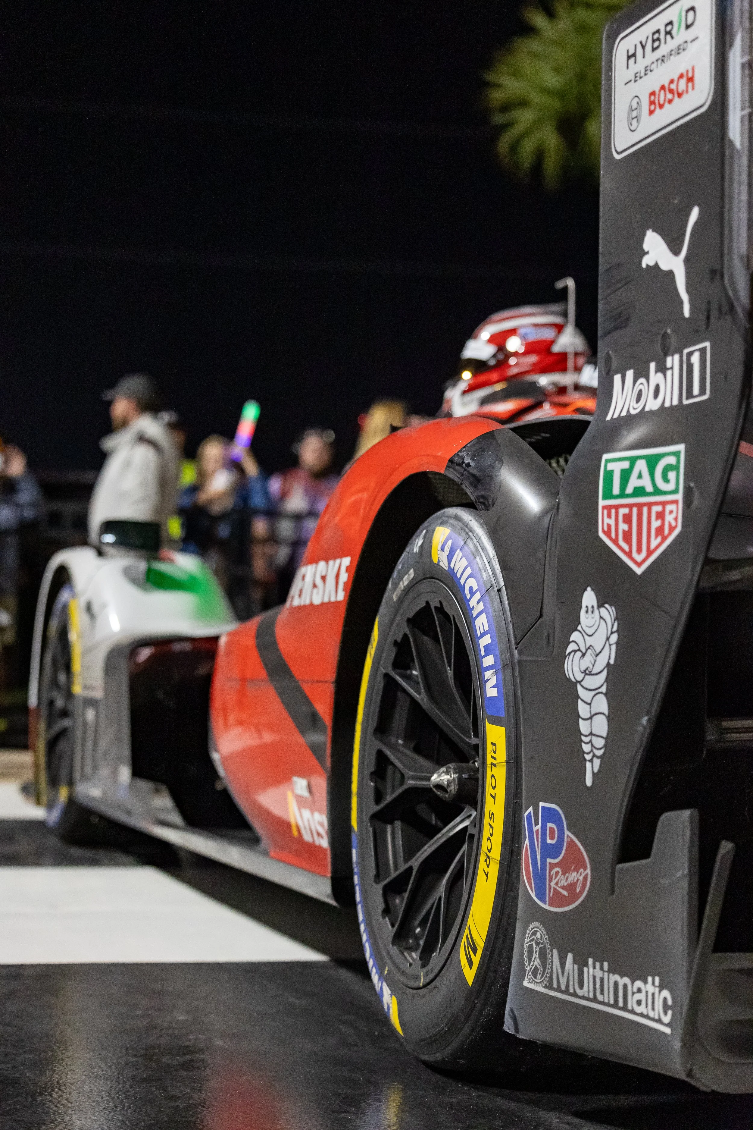 Close-up of a race car with various sponsor stickers, including Bosch, Mobil 1, TAG Heuer, and Michelin, on a black background. The car has a red, black, and white color scheme, and a helmet is visible toward the back. A group of people is blurred in