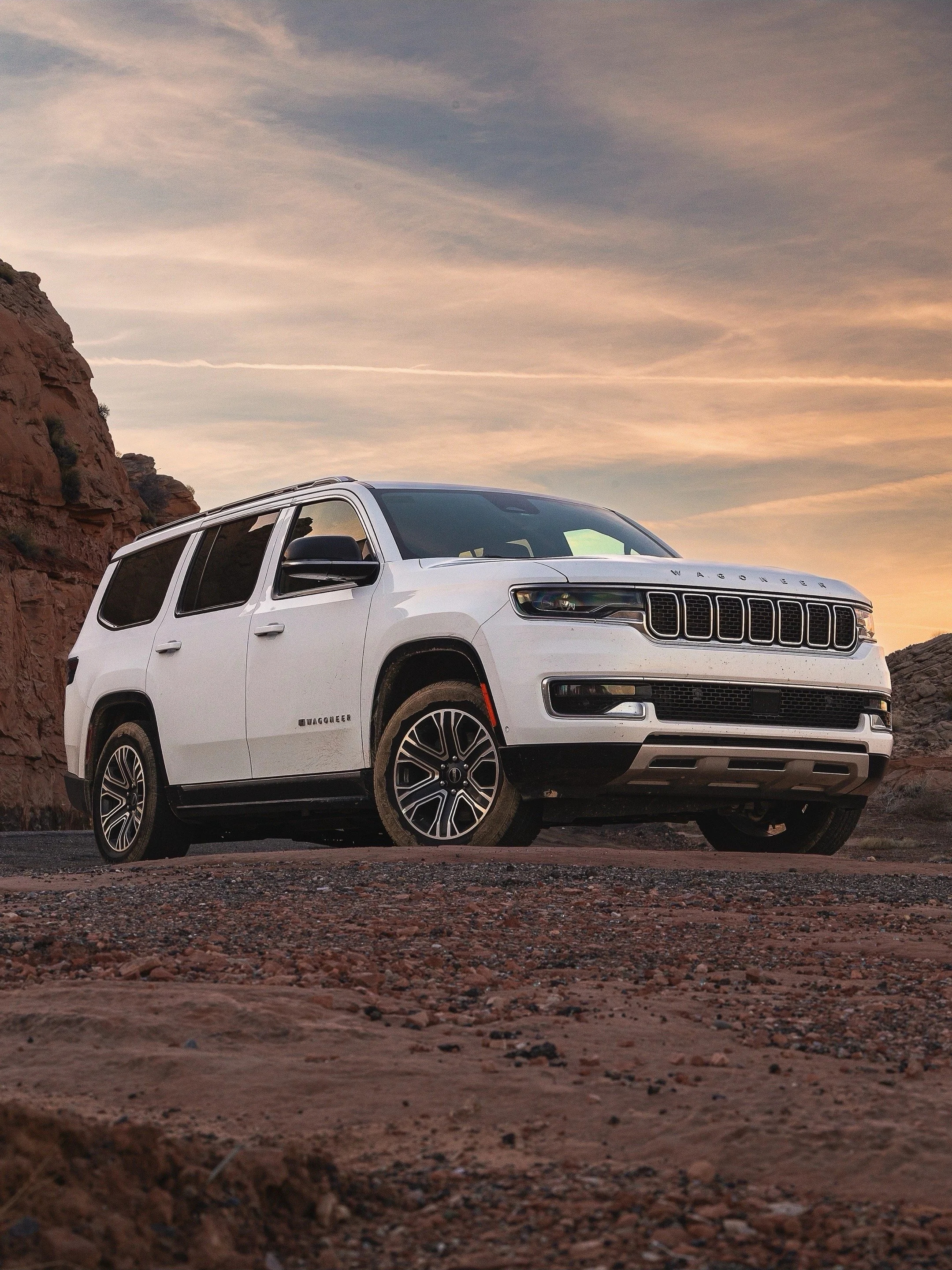 A white Jeep Wagoneer parked on rough dirt terrain with rocky hills and a sunset sky in the background.