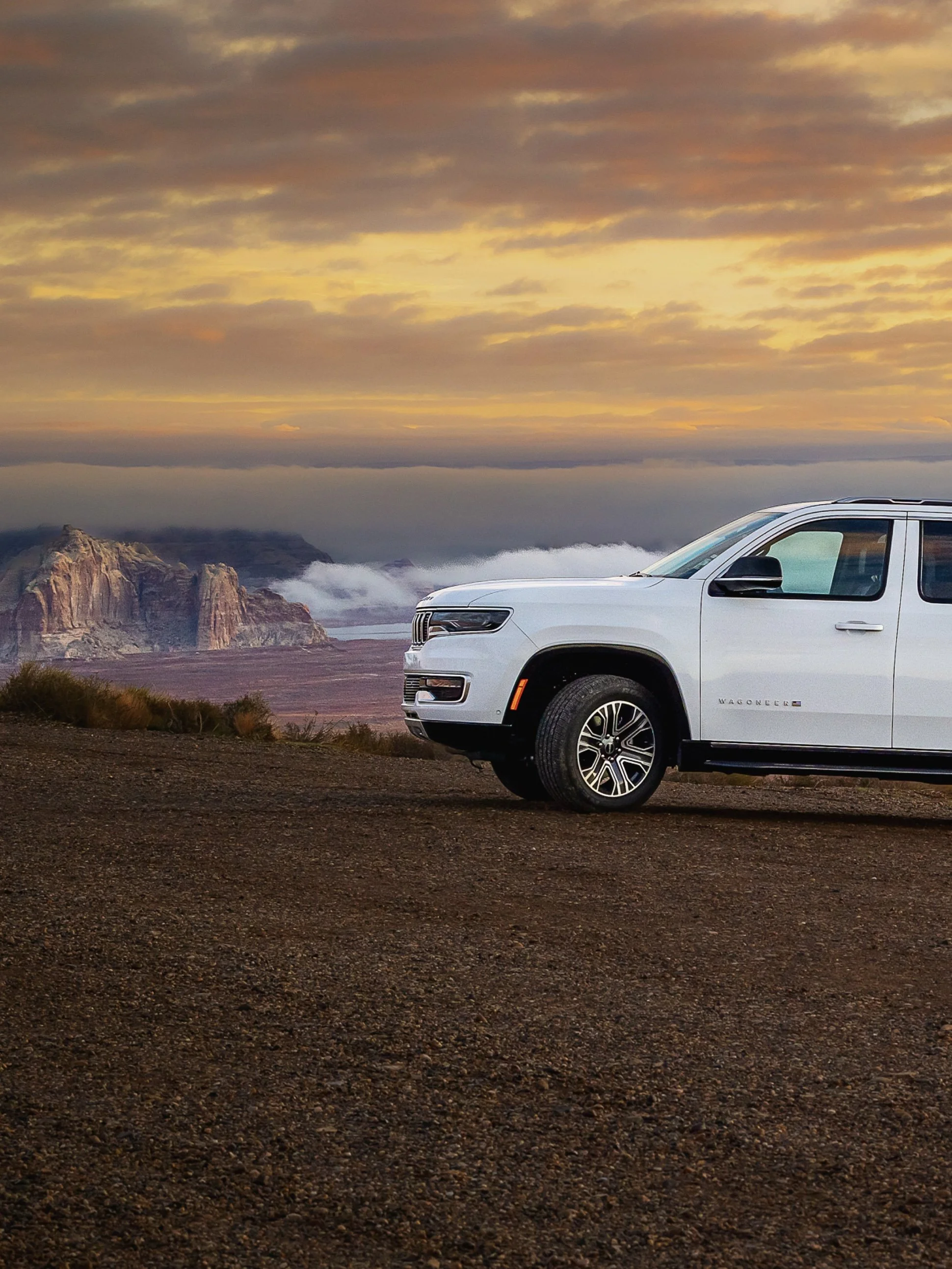 A white SUV parked on a dirt ground with a mountainous landscape and a colorful sunset sky in the background.