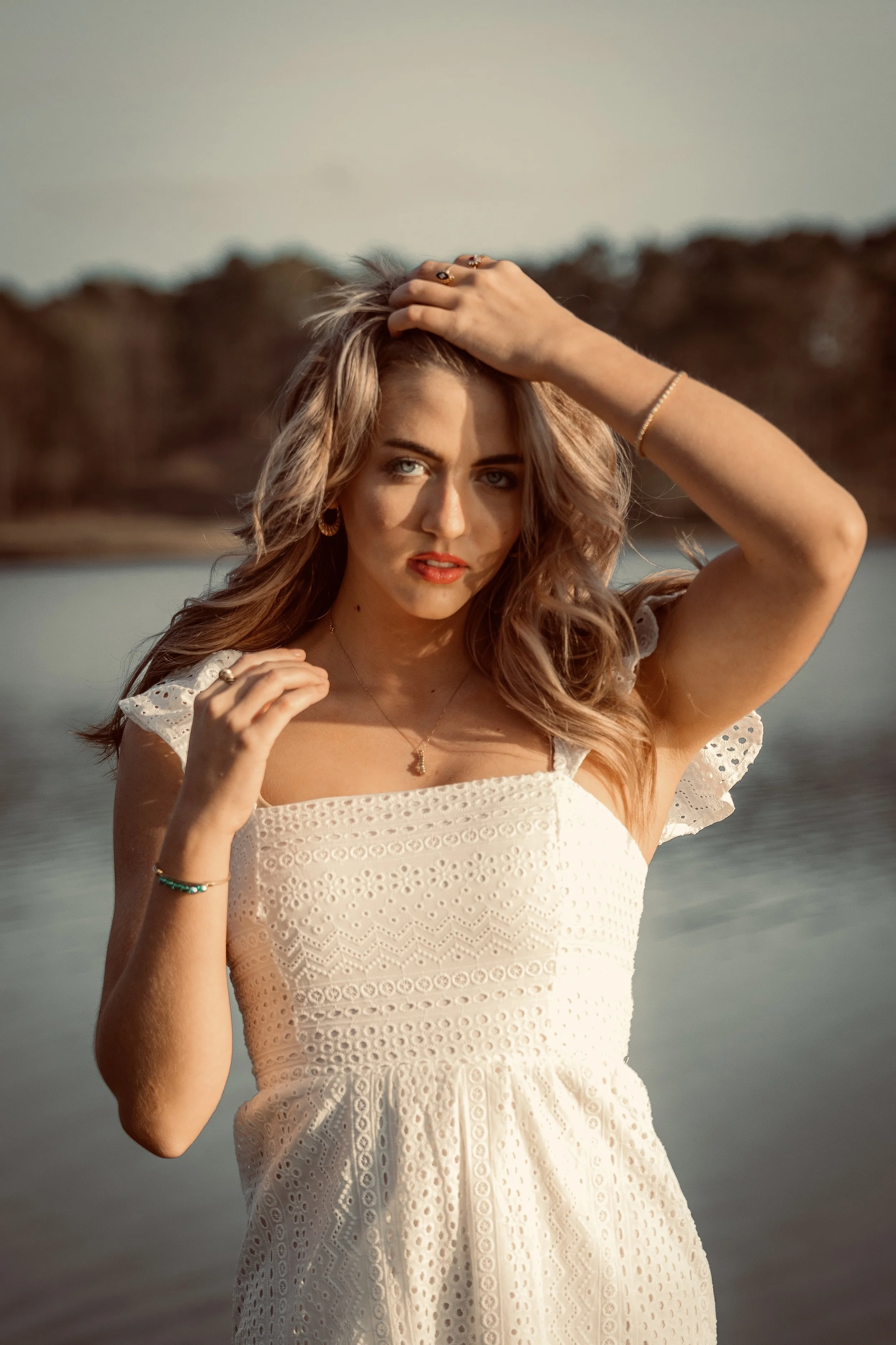 A woman with blonde hair and blue eyes wearing a white eyelet dress, holding her hair with one hand, and standing near water with a blurred background of trees and hills.