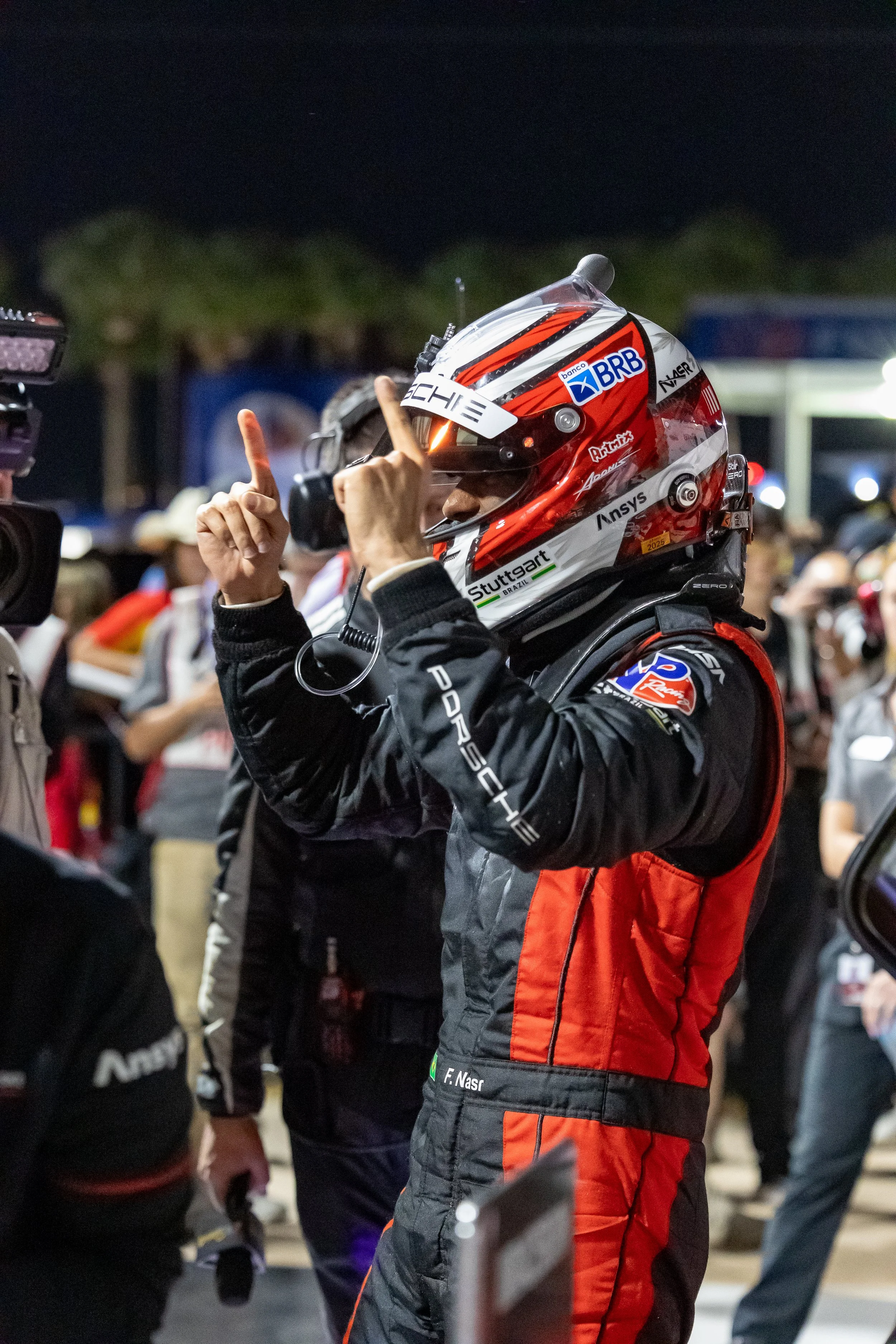 A race car driver with a helmet and racing suit talking to someone at a racing event during the night. The helmet has sponsor logos, and the suit is black with red and gray accents.