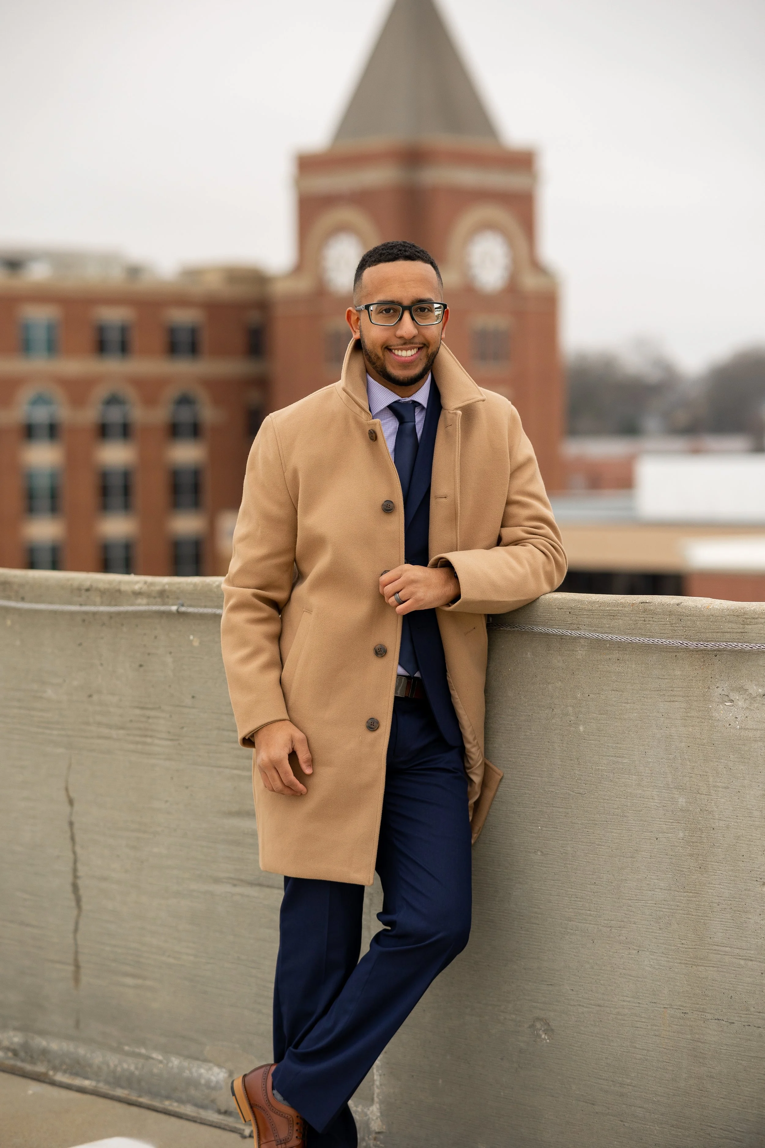 A man stands outdoors on a rooftop, wearing glasses, a tan coat, a blue shirt, and dark pants, smiling with a historic brick building with a clock tower in the background.