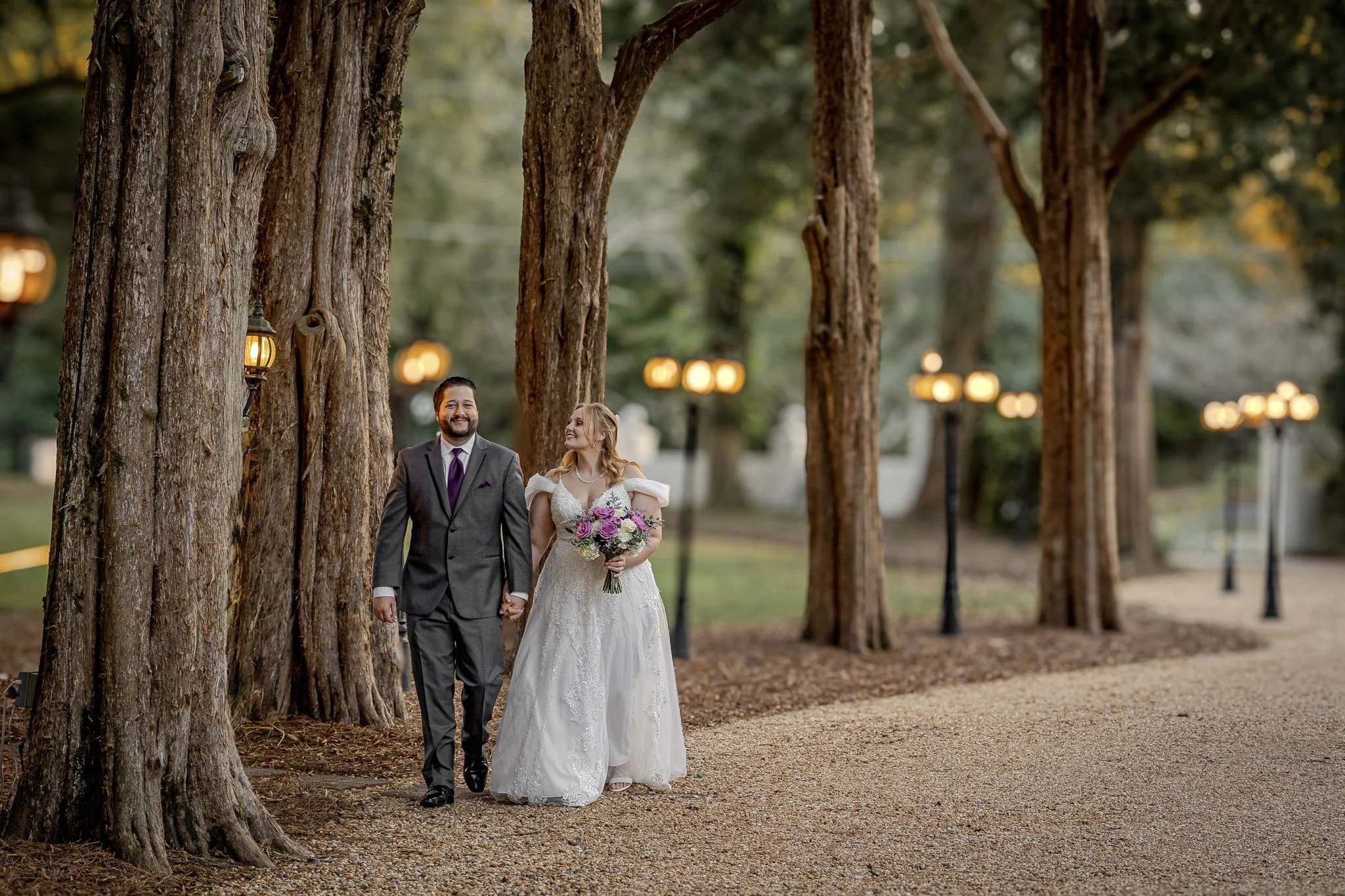A newlywed couple walking hand in hand through a grove of large trees, illuminated by warm streetlights, during sunset or twilight.