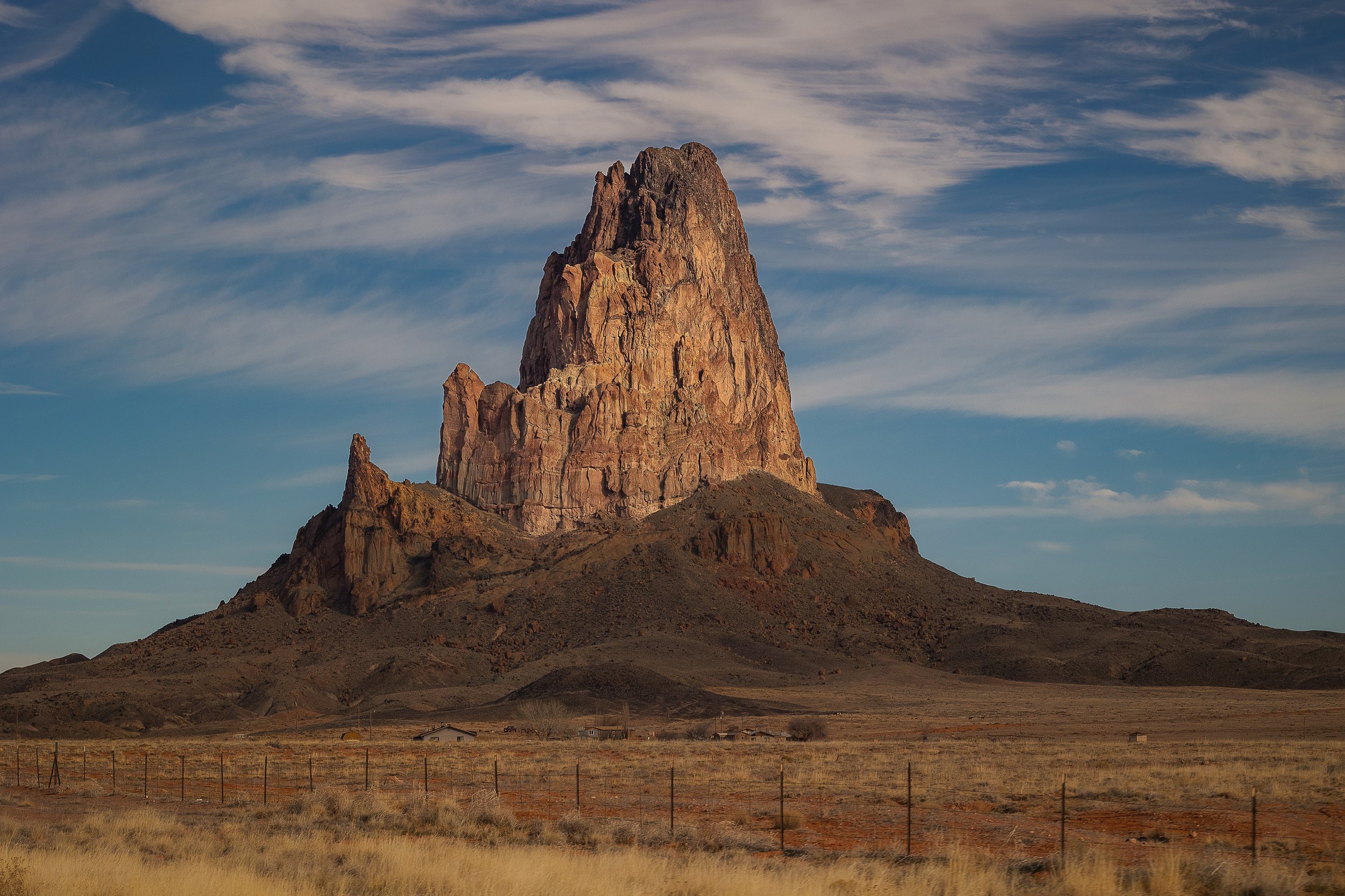 A large, rugged rock formation rises prominently from a flat desert landscape under a partly cloudy sky.