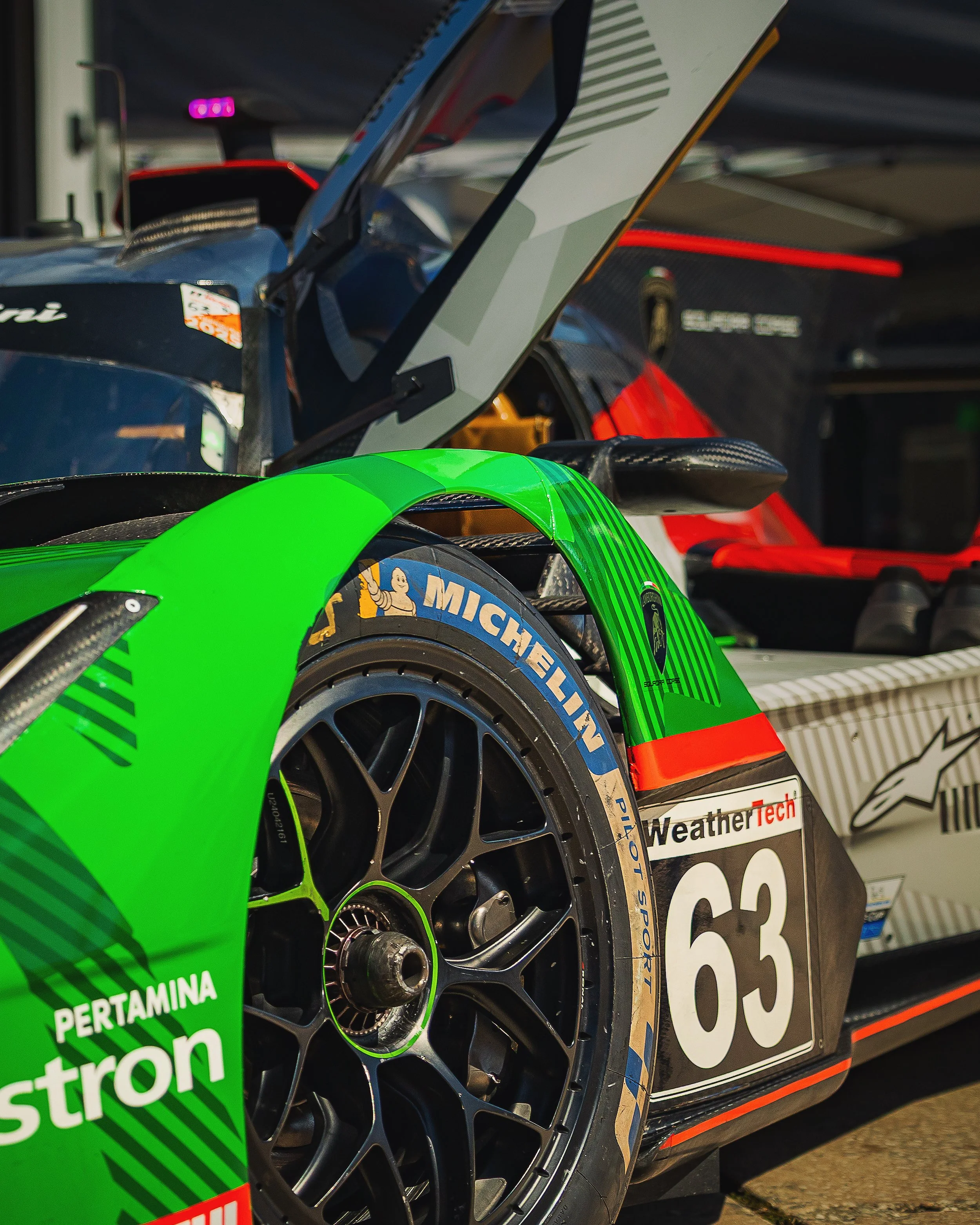Close-up of a race car wheel and tire with sponsor logos, carbon fiber components, and a partially visible card in the background.
