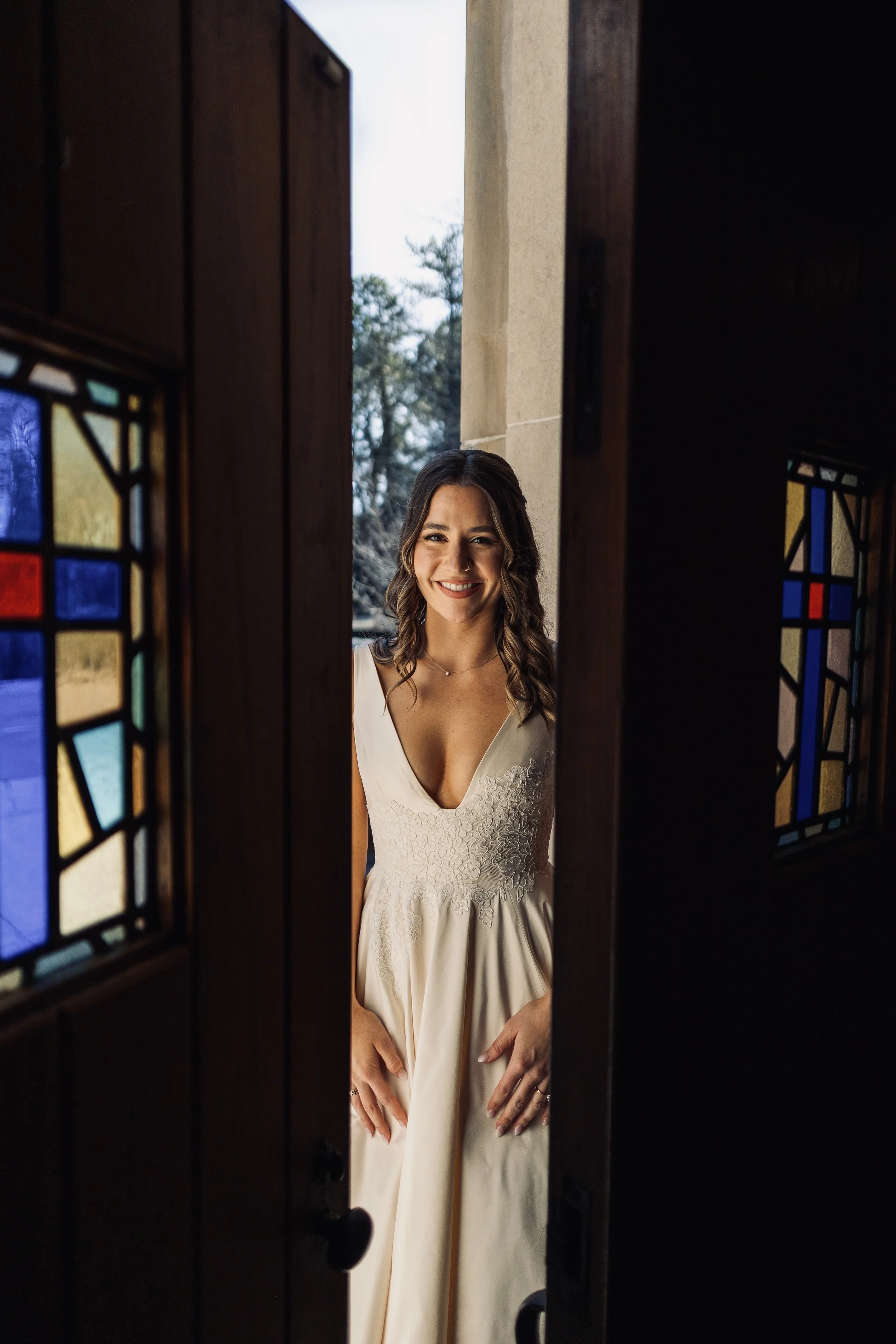 A woman in a white dress smiling through a partially open wooden door with stained glass panels.