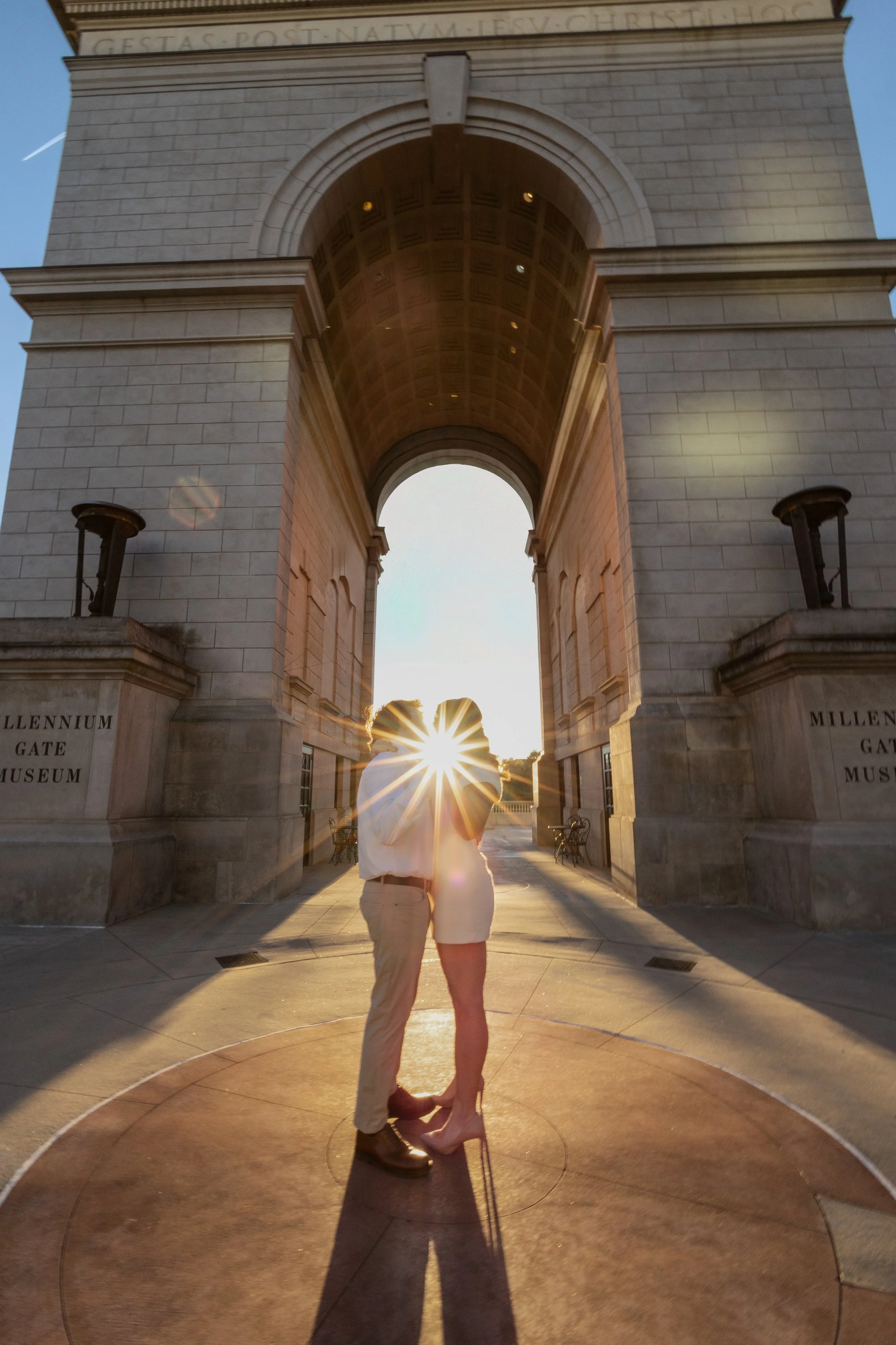 A couple stands kissing under an arch at sunrise or sunset, with a person’s silhouette and the sun visible through the arch, creating a starburst effect.