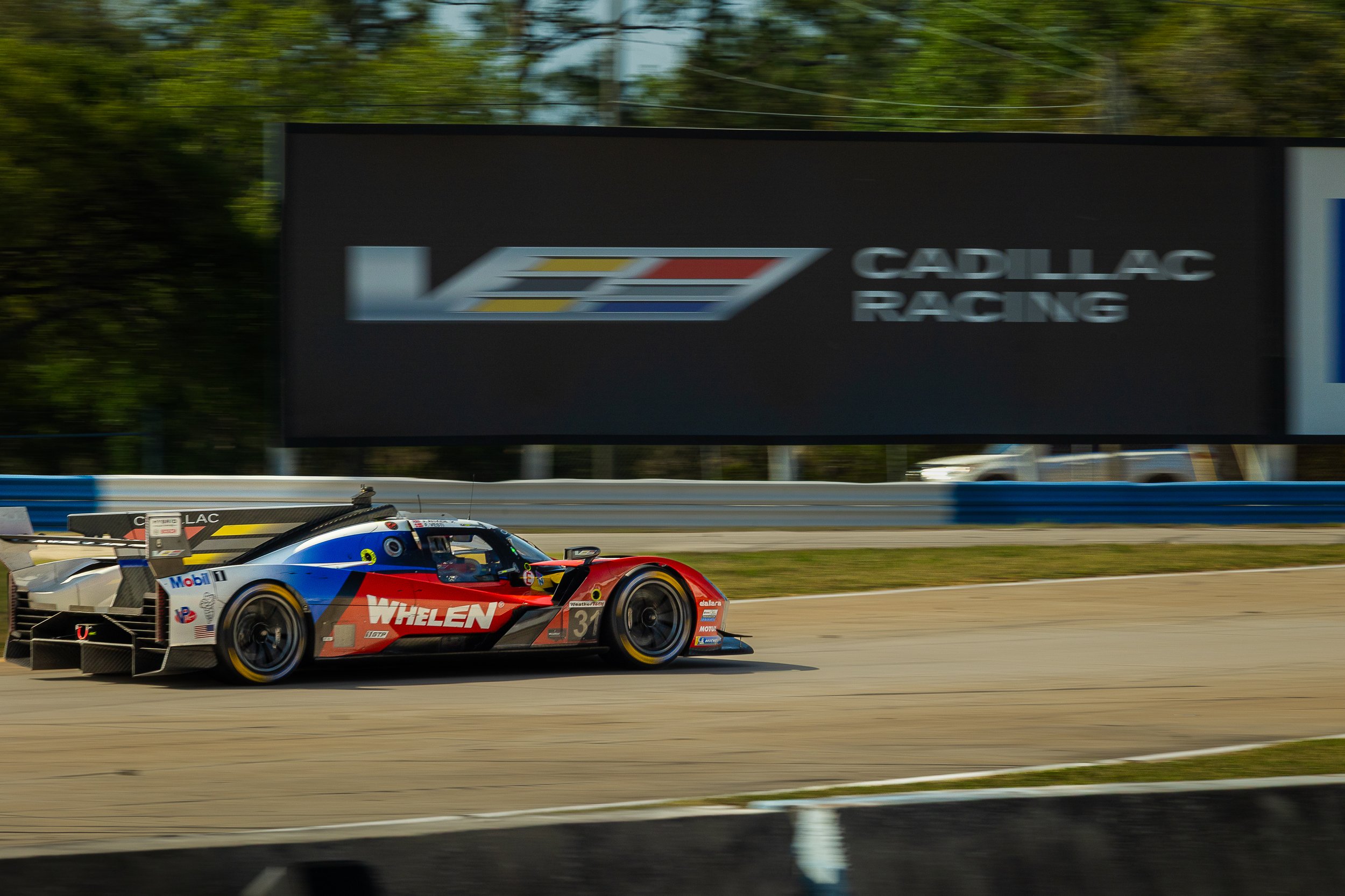 A race car with 'Whelen' branding speeding on a track with a large Cadillac Racing billboard in the background.
