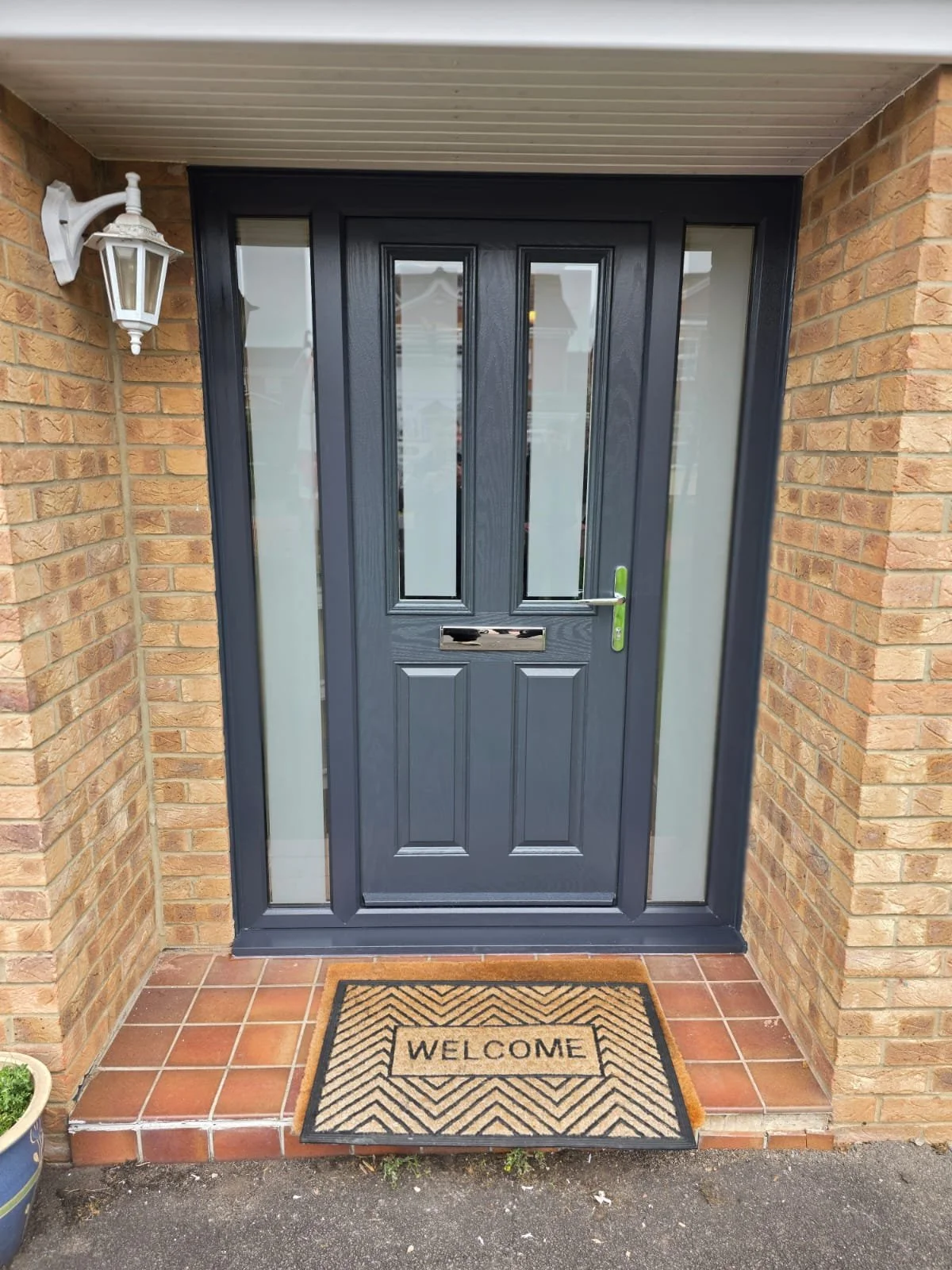 Front door with a dark gray finish, glass panels, and a green handle, surrounded by brick walls and a porch with a welcome doormat.