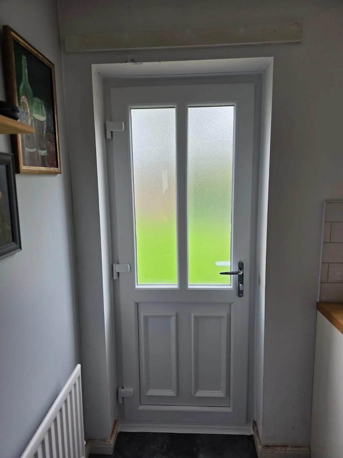 White exterior door with glass panels, handle, and lock, in a house interior with gray walls, a photo on the wall, a radiator below it, and a wooden countertop to the right.