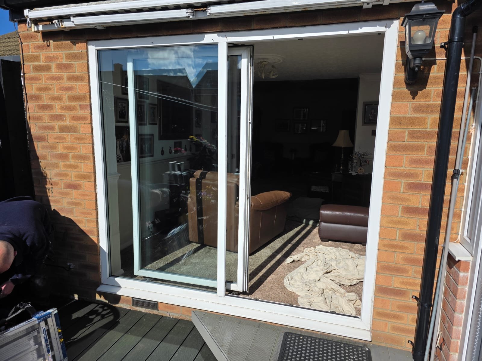 Sliding glass door leading to a living room with furniture inside, a lamp, and framed pictures on the wall. The exterior has a brick wall, outdoor lamp, and wooden deck flooring.