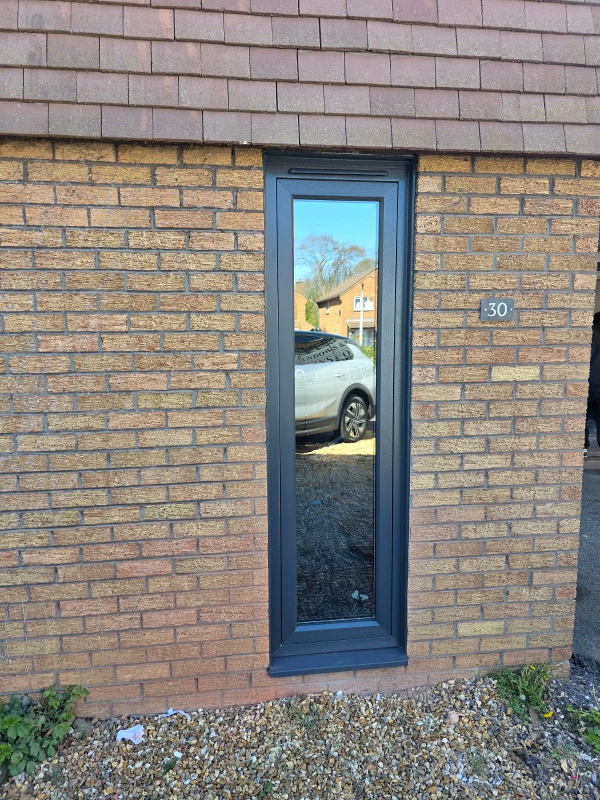 A narrow window set into a brick wall, reflecting a car and a house in the background.