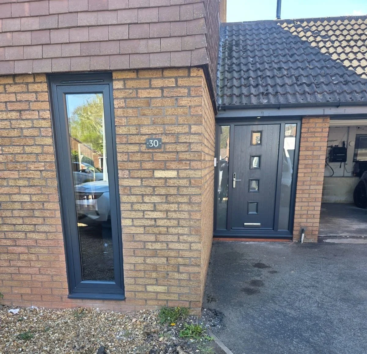 Close-up of the entrance to a house with a modern exterior, featuring a black door with four small square windows, a silver handle, and a house number 30 on the brick wall. The house has brick walls and a sloped tiled roof. A narrow window reflects a parked car and trees, and there is an area of gravel and pavement in front of the entrance.
