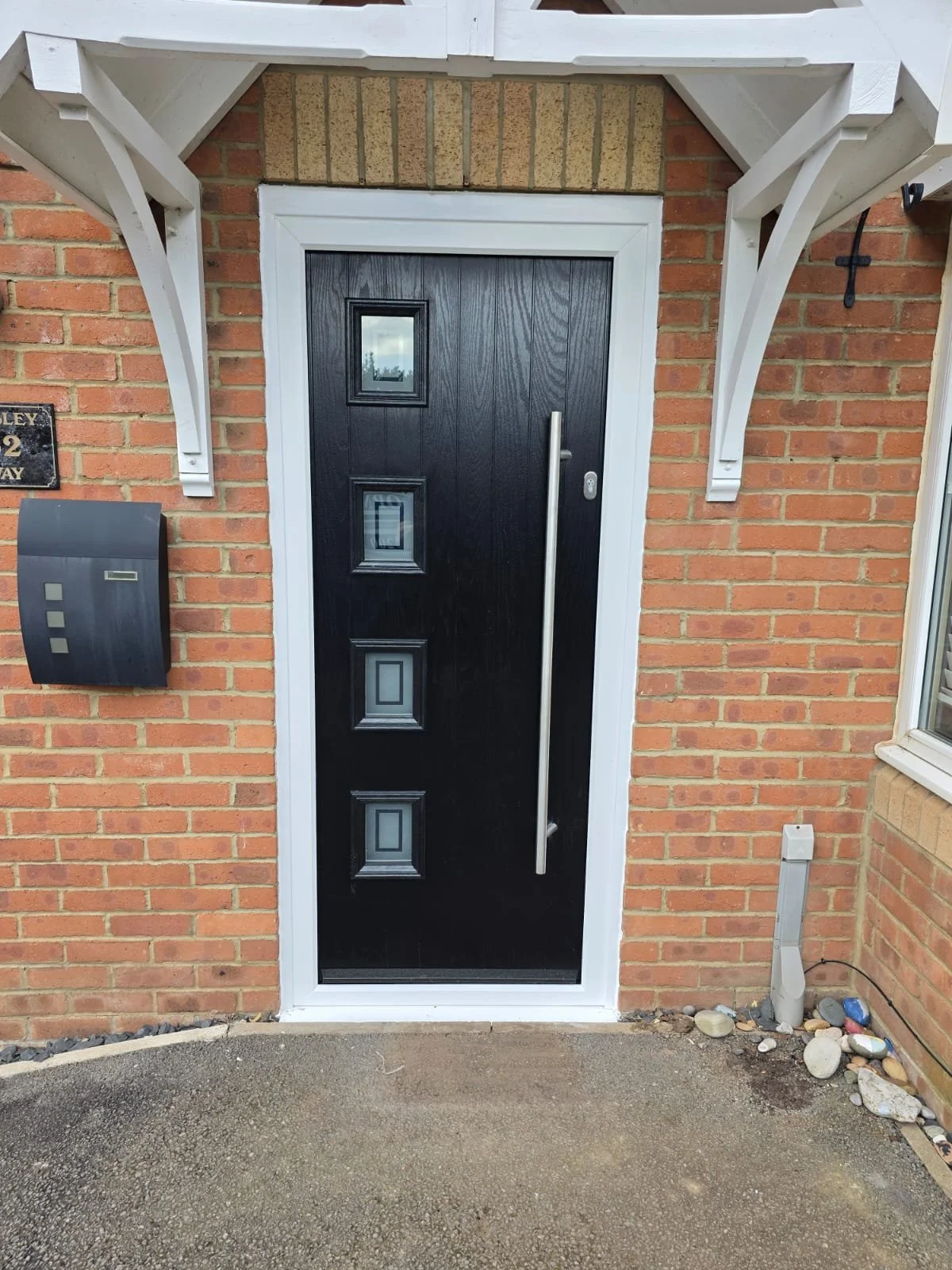 Black front door with three narrow decorative glass panels, white framing, and a long vertical door handle, set in a red brick house with a small porch, mailbox, and stones nearby.