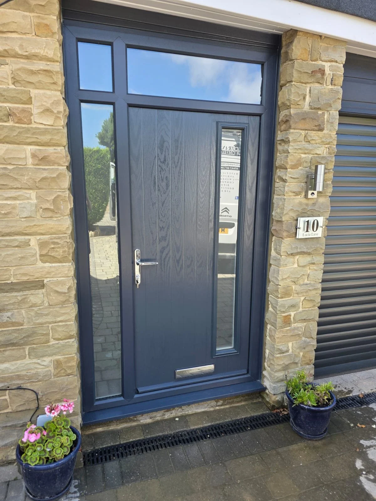 A modern front door made of dark gray wood with a vertical narrow window on the right, a horizontal window above, silver handle, and letterbox at the bottom. The door is set in a stone wall with a house number sign '10 Lucia Lane' on the right. There are potted plants with colorful flowers on either side of the door and a metal drainage grate on the ground in front.