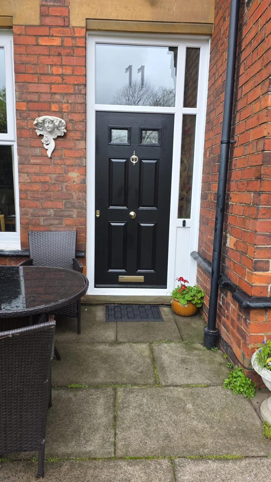 Front entrance of a brick house with a black door, a decorative wall sculpture of a woman's face, a house number 11, a small outdoor table with chairs, and potted plants.