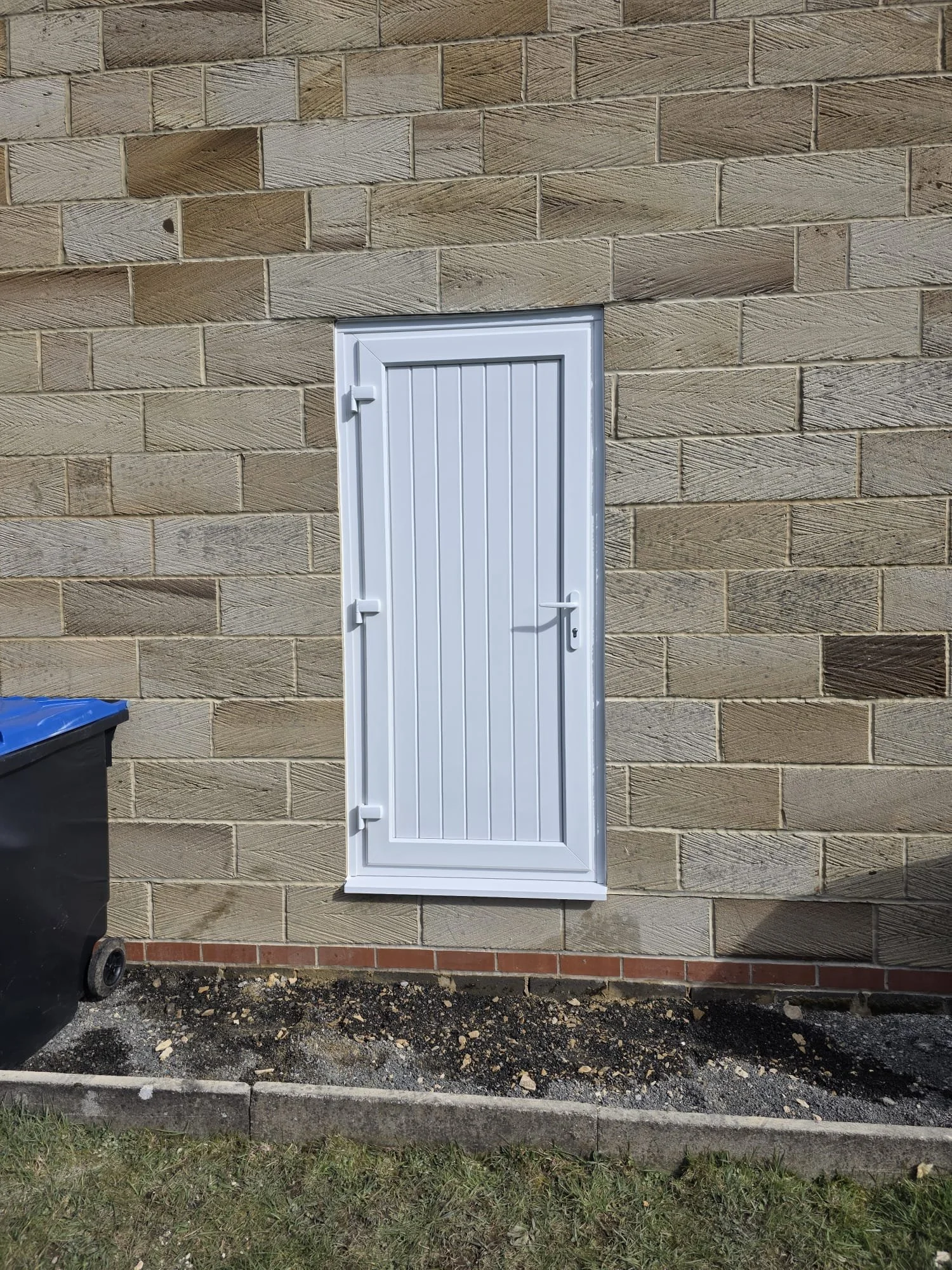 A white metal door with vertical slats on a brick exterior wall. There is a black trash bin with a blue lid on the left side and some dirt and grass at the bottom edge of the image.