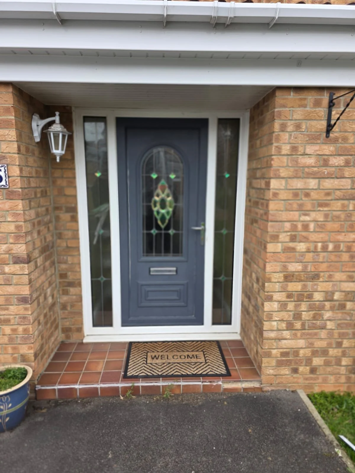 Brick house front entrance with a dark gray door featuring decorative glass panels, a black and beige welcome mat, a potted plant, and exterior lighting on the brick wall.