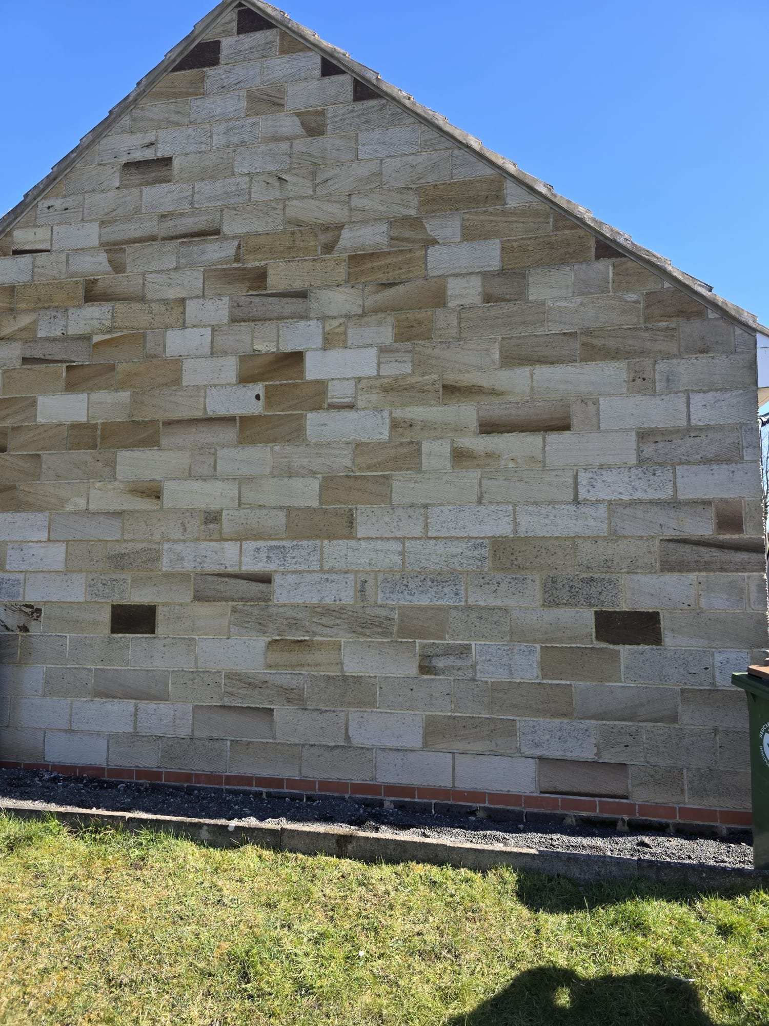 A building with a wall made of unfinished wood or stone blocks, with a gable roof, against a clear blue sky. The base of the wall has a red brick layer, and there is grass and a shadow in the foreground.