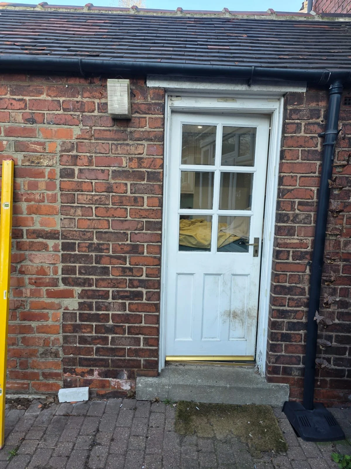 White door with glass panels on a brick exterior wall, with a step, a yellow level on the left, a black pipe running down the right side, and a vent on the wall above the door.