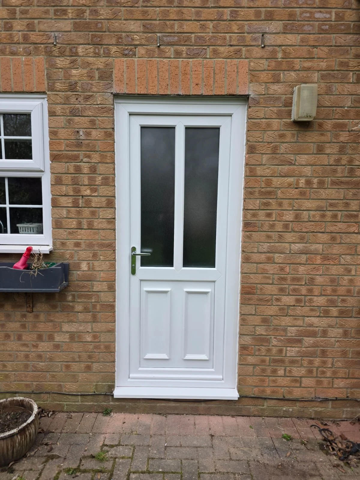 White door with frosted glass panels on a brick wall exterior of a house. There is a window with white framing to the left of the door, a flower box with gardening tools beneath, and an outdoor light fixture to the right of the door. The ground is pa