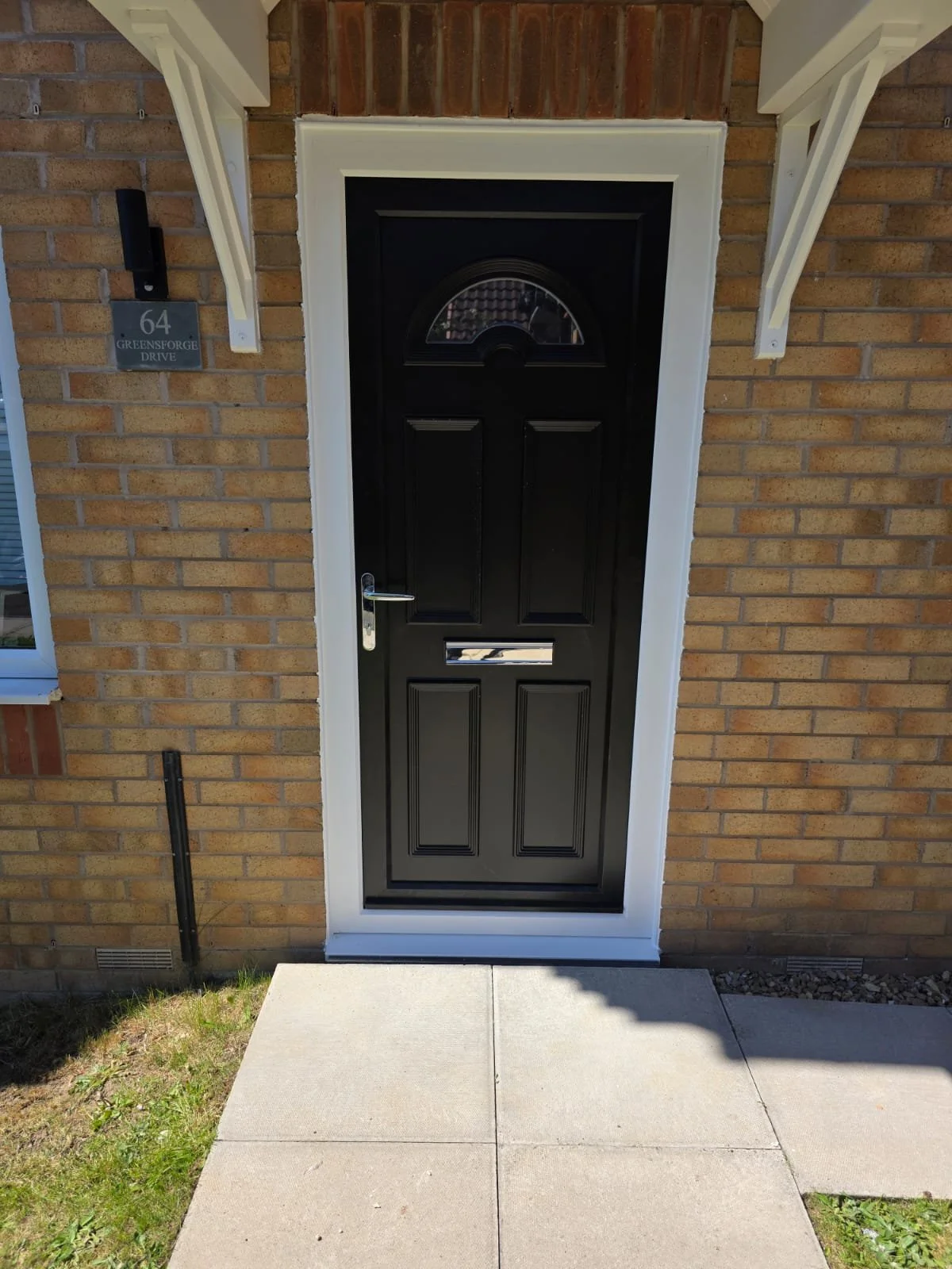 Black front door with window at the top, surrounded by tan brick wall, with a silver handle and mail slot. White trim and porch supports, house address 64 Greensforge Drive sign to the left.