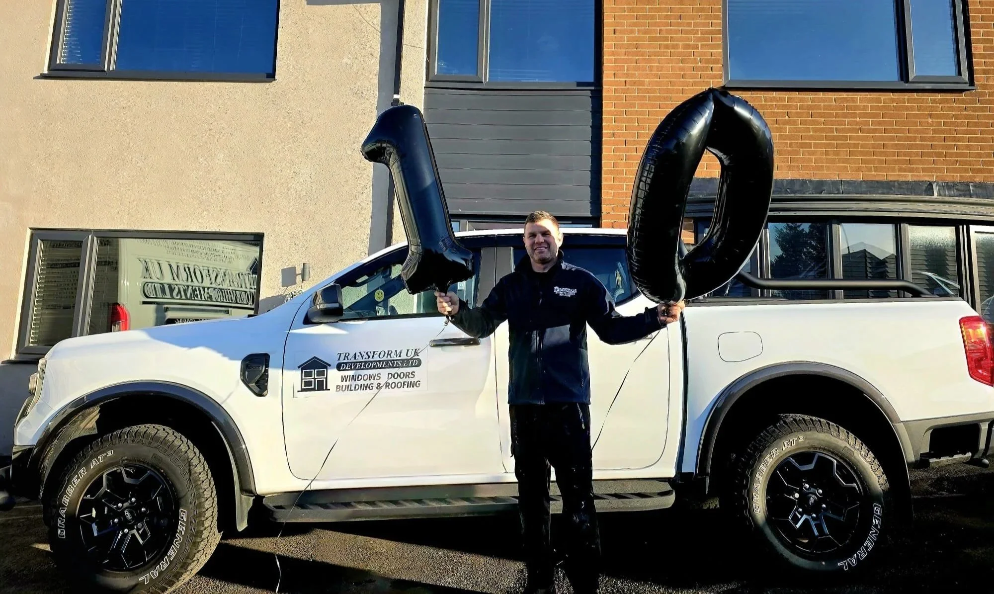 A man standing in front of a white pickup truck holding two large black helium balloons shaped like the number two and the number zero. The man is smiling, wearing a black jacket, and the background shows building windows and a partly cloudy sky.