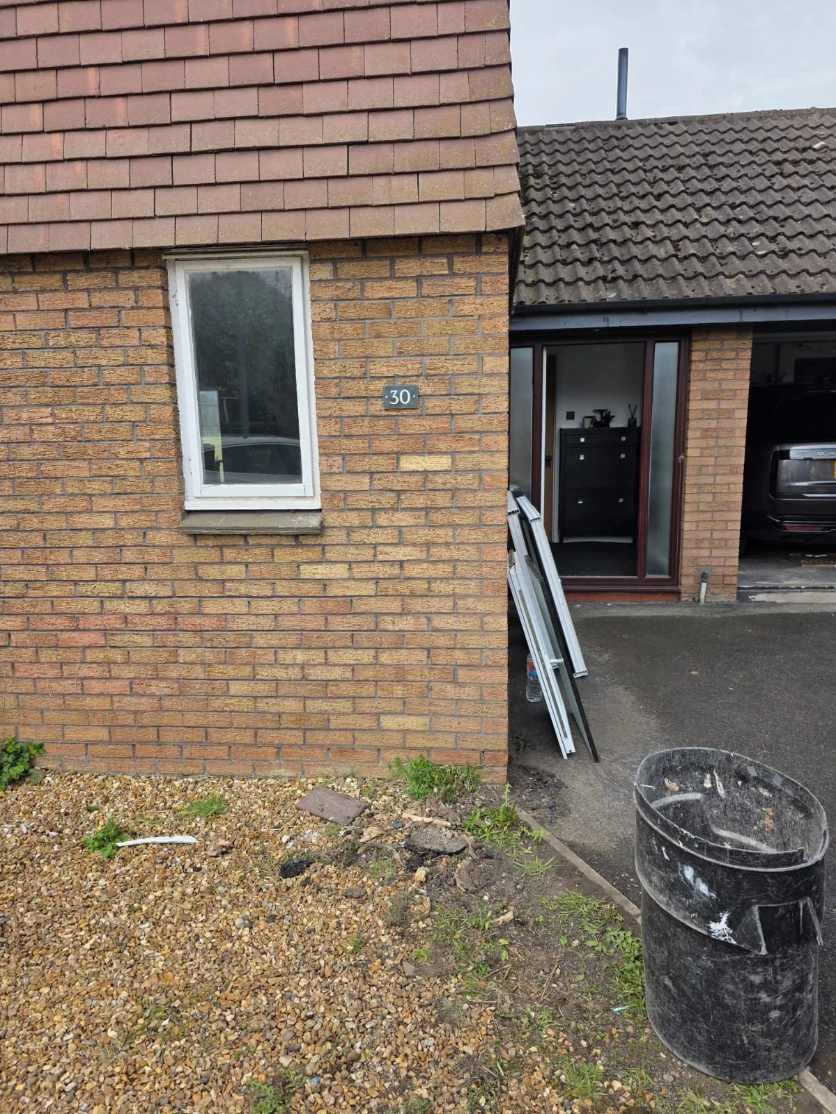 Exterior view of a house with a brick wall and garage. There is a window on the brick wall with a house number 30. The garage door is open, showing a black car inside. Construction materials and tools are leaning against the wall near the garage, and