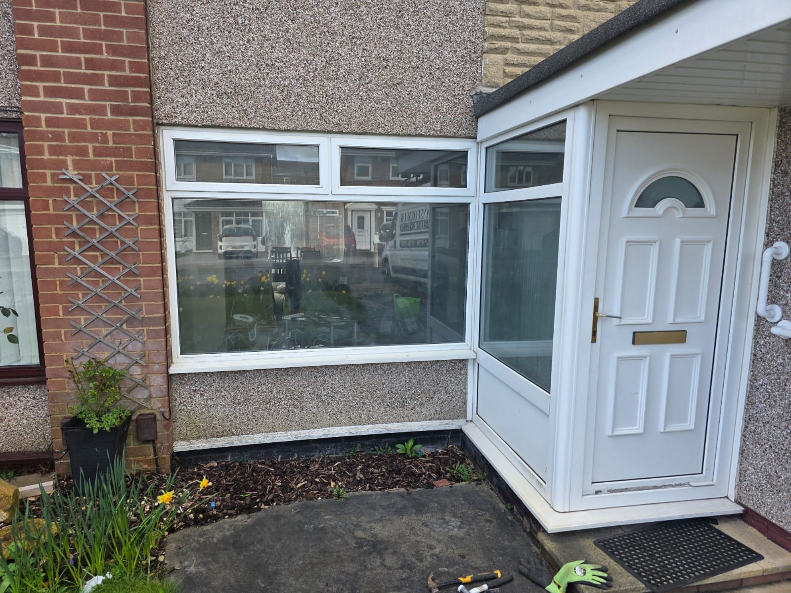 Front view of a house with a white enclosed porch, a white door with a semi-circular window, large front window, brick wall, potted plant, garden bed, gardening tools, and a doormat.