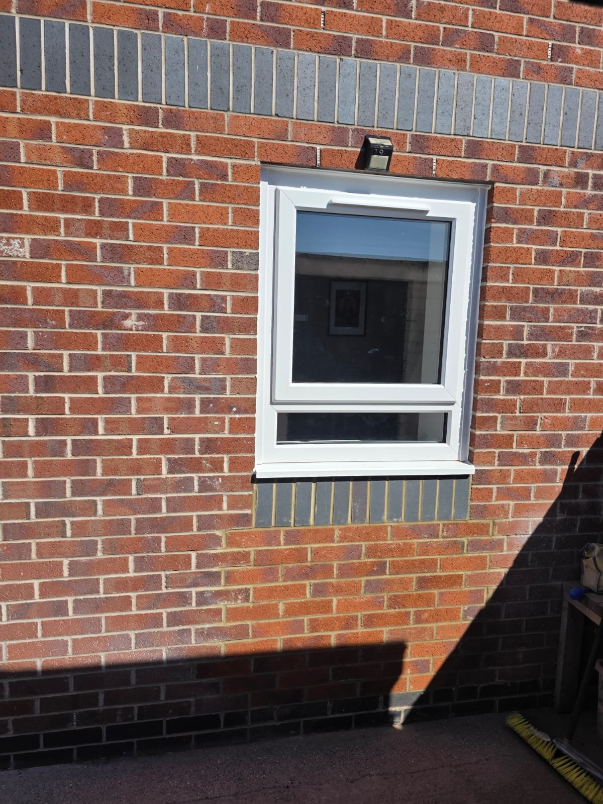 A white-framed window set in a brick wall, with a small black device mounted above it and a gray vent below, sunlight casting a shadow on the ground.