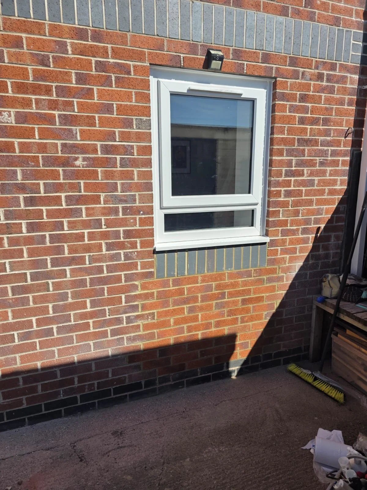 A brick building exterior with a white-framed window. There is a small black security camera above the window. The ground in front of the building has some tools and debris.