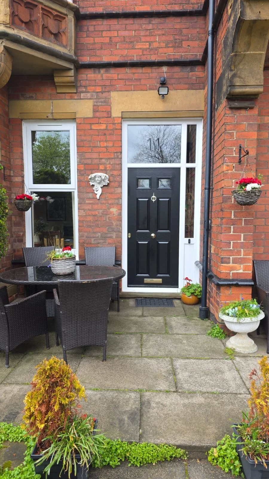 Front porch of a house with a black door, two windows, hanging flower baskets, potted plants, a patio table with chairs, and decorative wall elements.