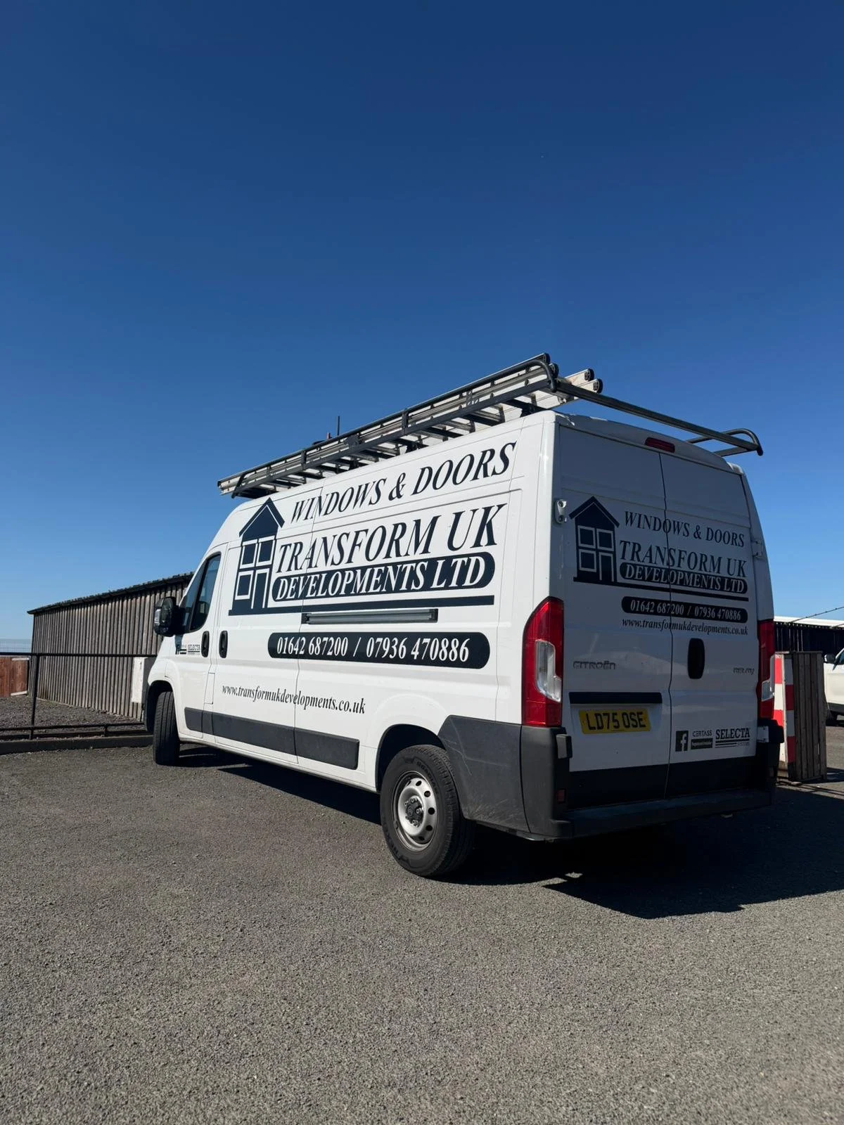 A white company van with black and blue logo and text advertising Windows & Doors and Transform UK Developments LTD, parked on a gravel surface under a clear blue sky with a wooden fence in the background.
