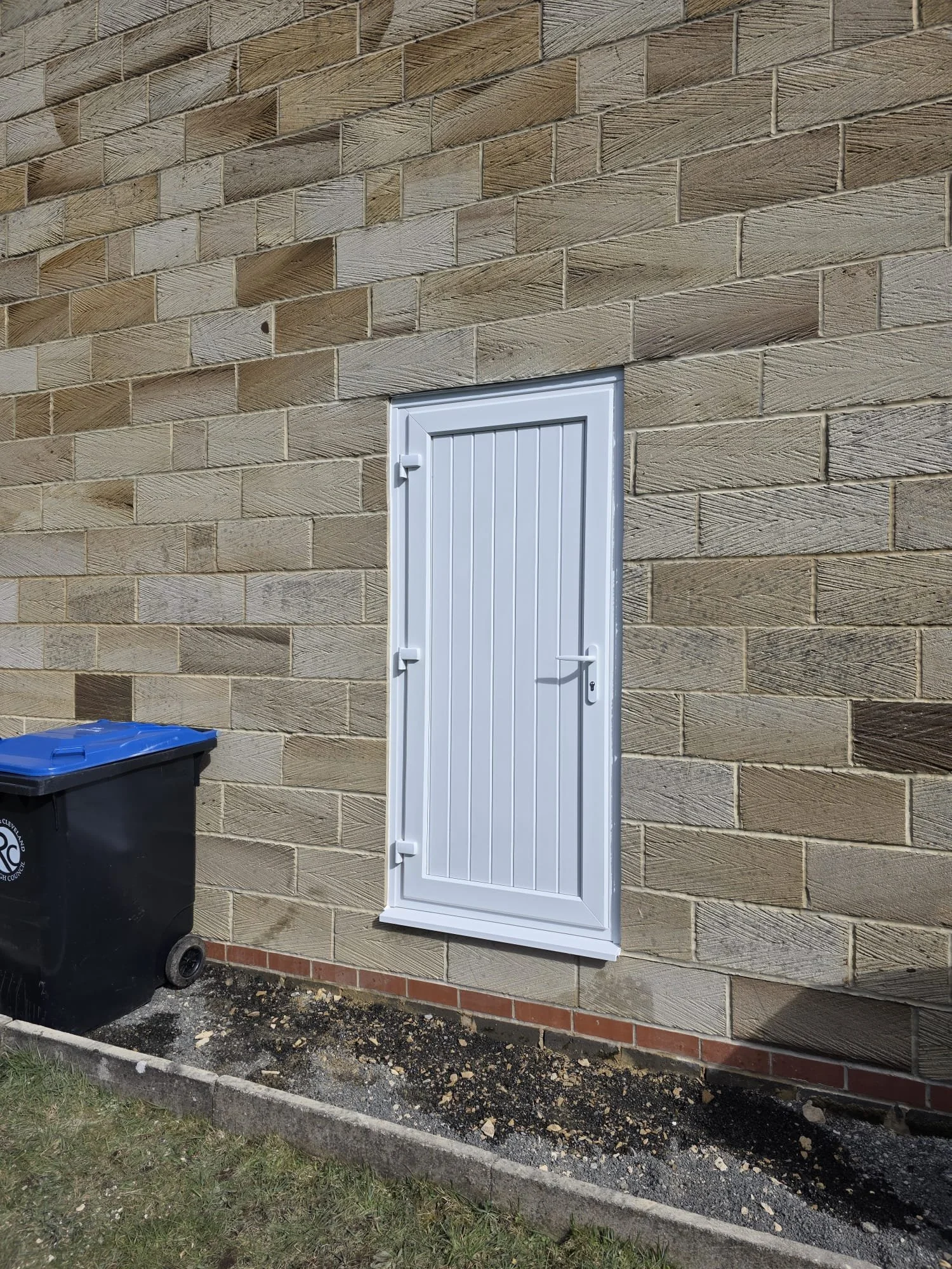 A white outdoor utility door mounted on a brick wall. To the left, a black trash bin with a blue lid is placed on a small patch of grass, with a dirt and gravel area in front of it. The brick wall has an uneven pattern with some bricks protruding.