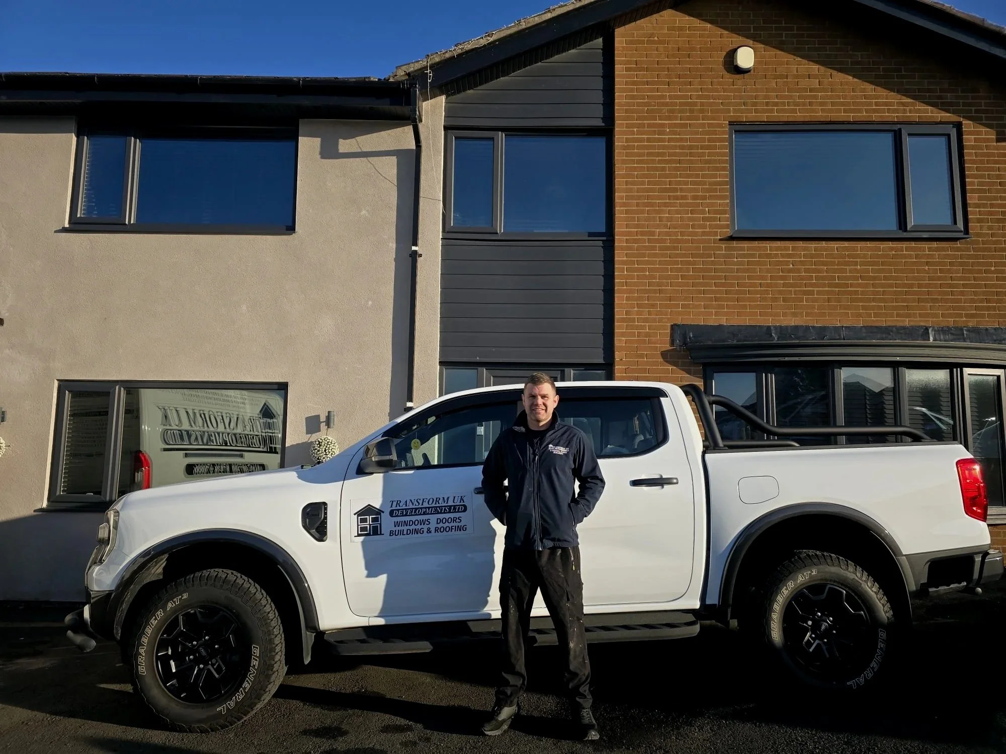 A man stands in front of a white pickup truck with company branding, parked in front of modern residential buildings with different exterior finishes. The man is wearing a black jacket and black pants.