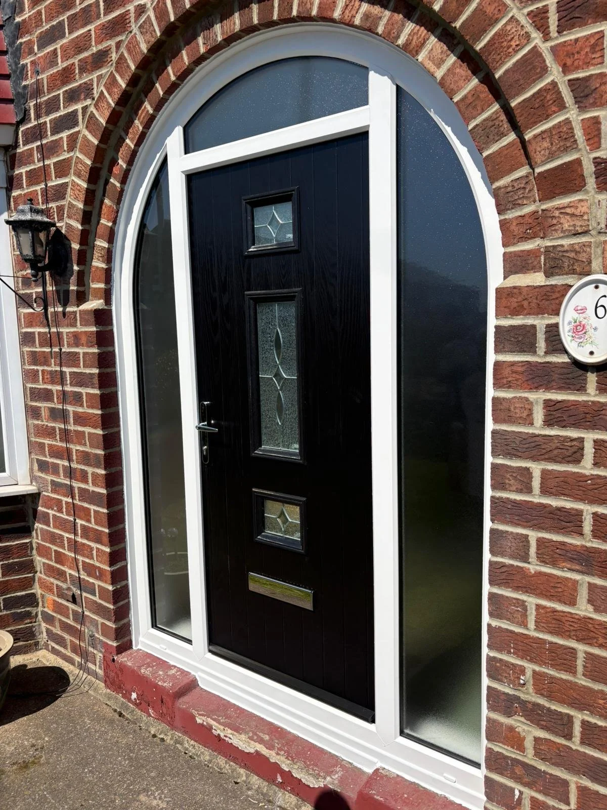 Front porch of a brick house with a black front door featuring decorative glass panels, surrounded by a white frame, and a black outdoor wall lantern on the left side.