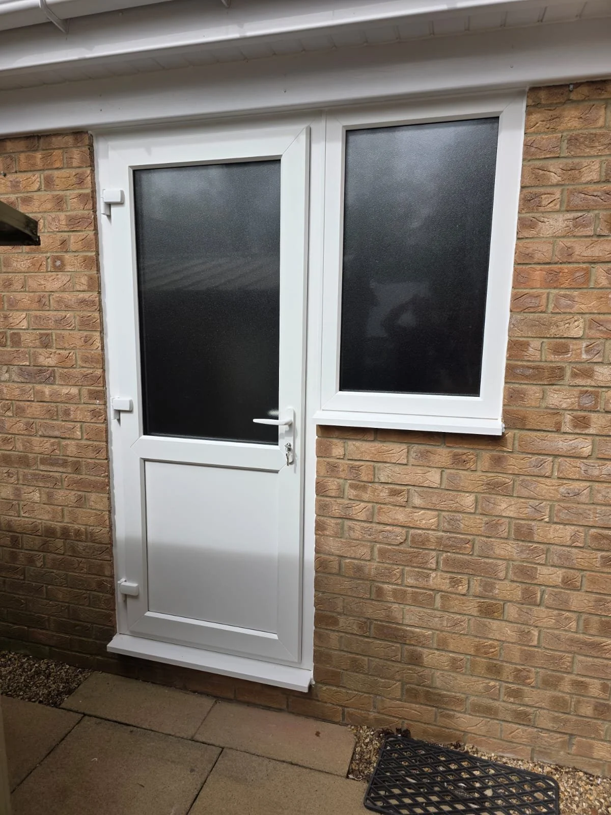 White door with a frosted glass window next to a window on brick wall, in an outdoor setting with paving stones and a black grate.
